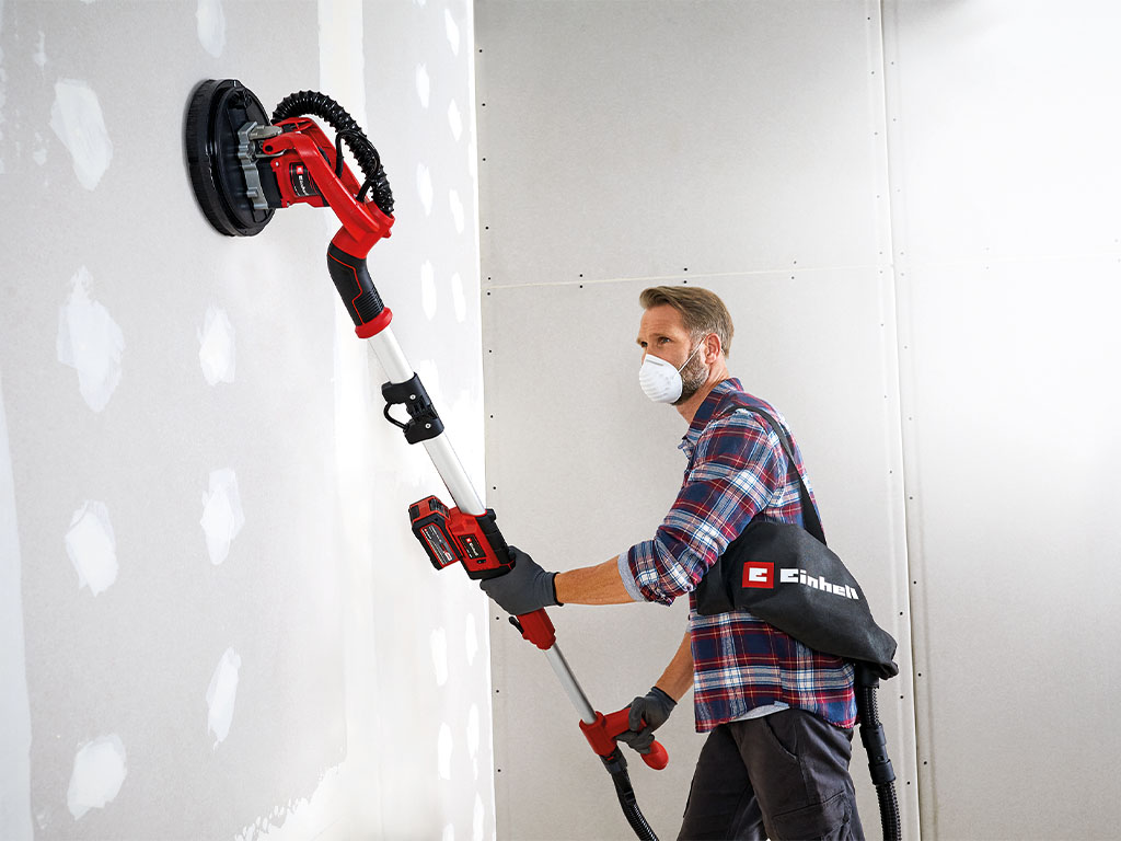 A man sanding drywall with an Einhell cordless drywall sander, its dust bag hanging over his shoulder.